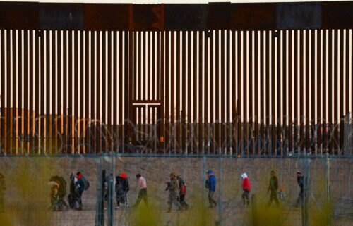 Migrants seeking asylum in the United States gather near the border wall after crossing a razor wire fence as a member of the Texas National Guard escorts them on December 19