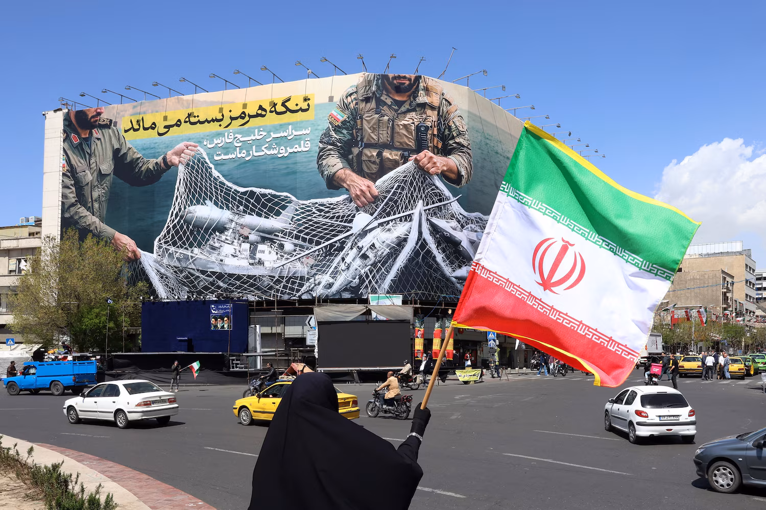 A woman holds Iran's national flag while standing near a billboard that reads "The Strait of Hormuz remains closed" at the Enqelab Square in Tehran, on April 5, 2026.

