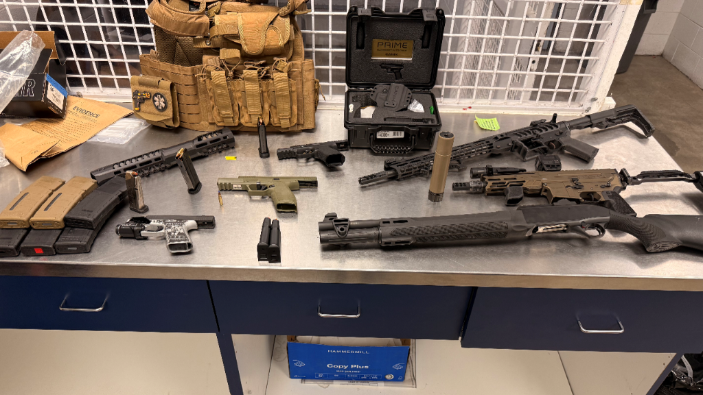 A workbench with various disassembled firearm parts, magazines, and a tactical vest arranged on the surface.